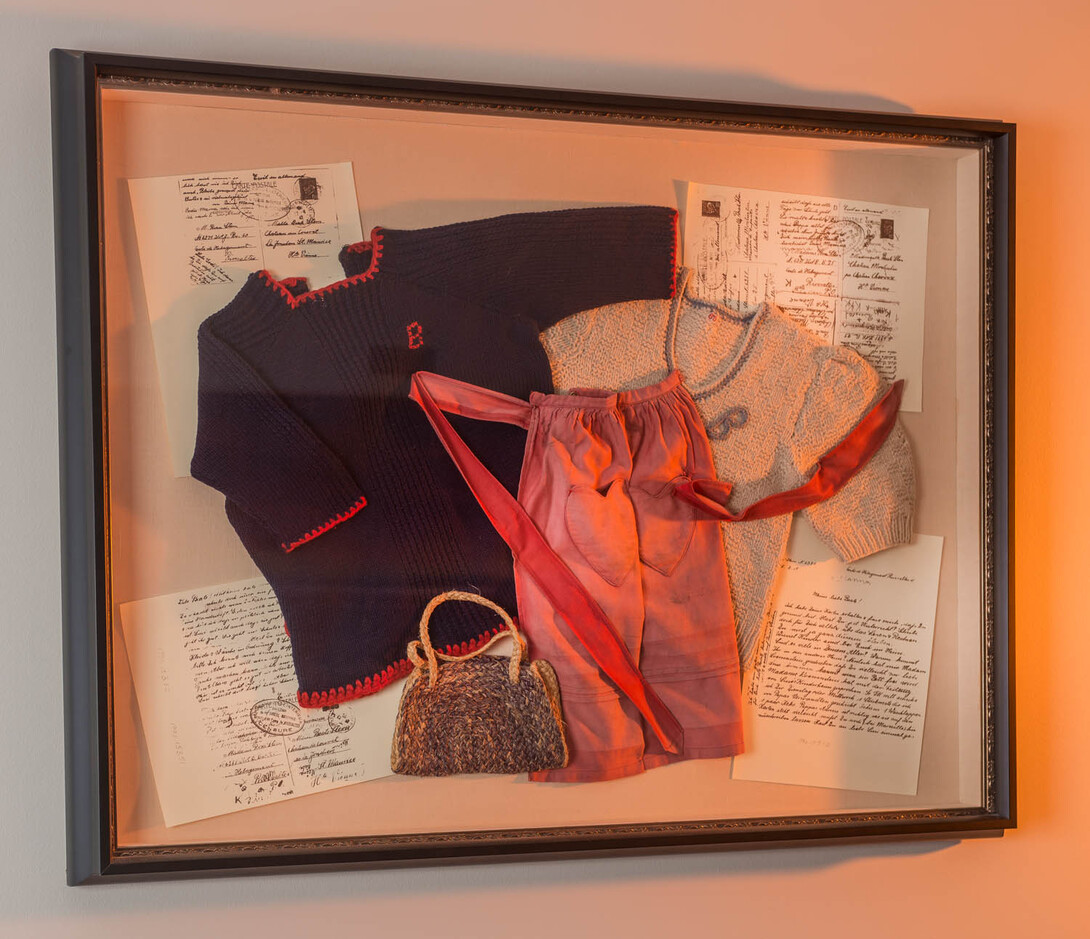 Framed display of vintage clothes, handwritten letters, and a small woven bag.
