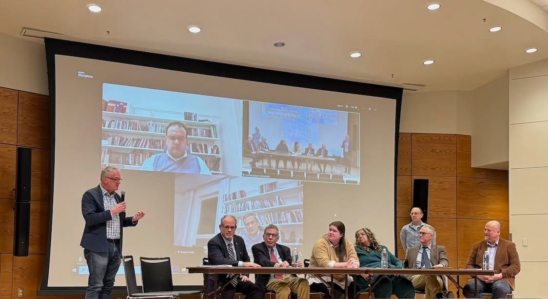 Panel discussion with six seated participants and one standing speaker in a conference room. Screen displays video call.