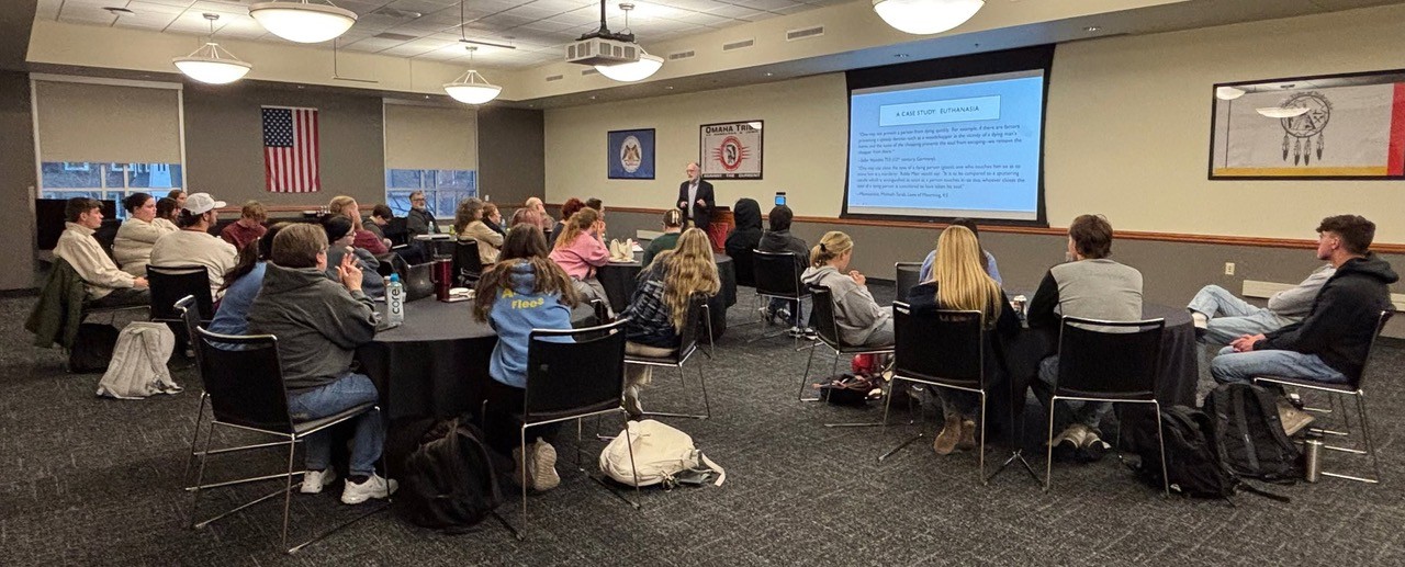Classroom setting with students seated at tables, facing a presenter and a projected slide.